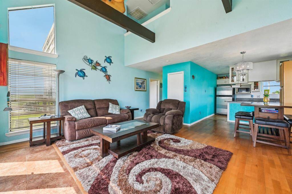 Bright living room with high ceilings, exposed beams, and large windows offering natural light and outdoor views. Features a cozy fireplace, modern furniture, and a wall-mounted TV, creating a welcoming atmosphere.