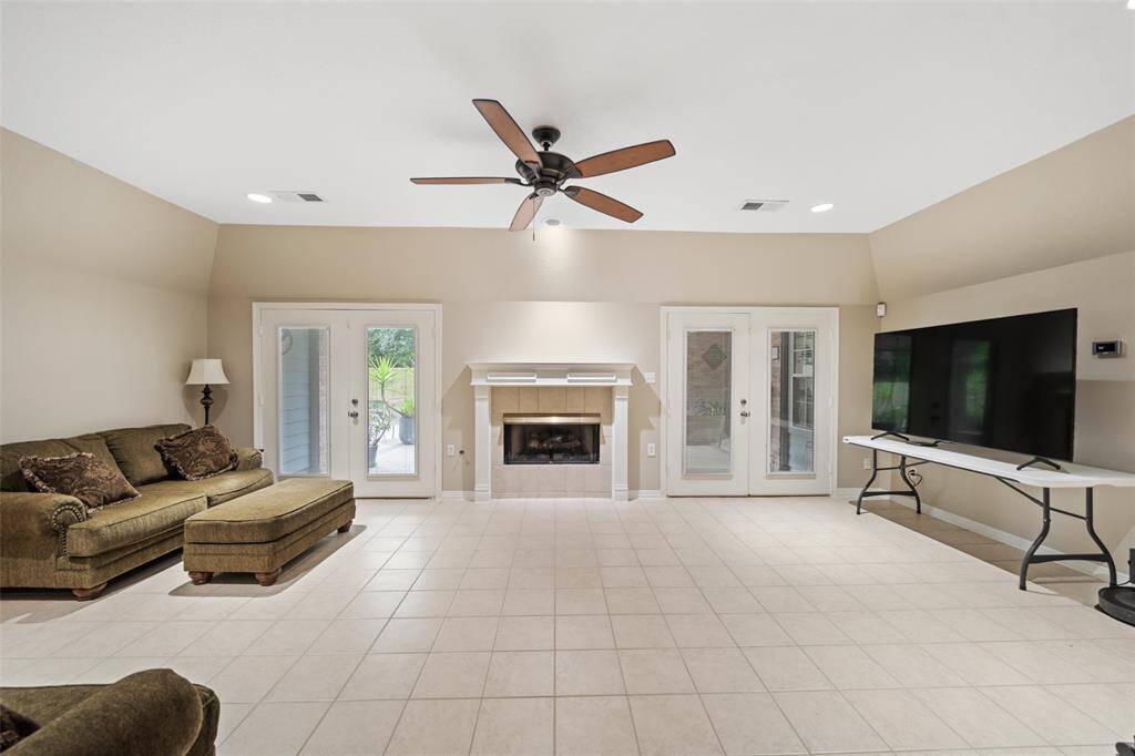 This spacious family room offers a warm and welcoming setting with tile flooring, soft neutral paint, a cooling ceiling fan, and a cozy gas-log fireplace with a traditional mantel. Two sets of French doors provide easy access to the backyard, while recessed lighting keeps the space bright and inviting.