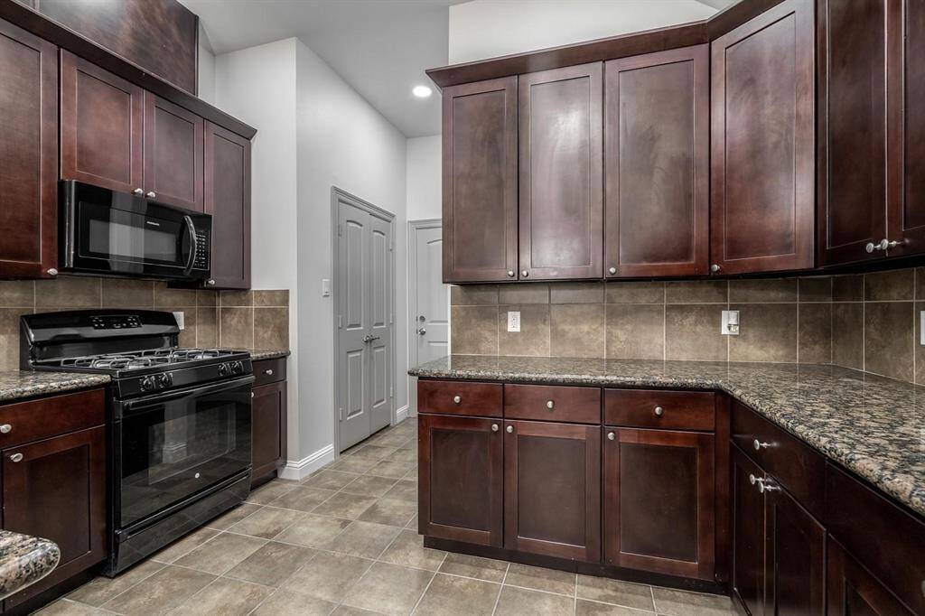 This kitchen features dark wood cabinets, a sleek black appliance suite including a gas stove and microwave, granite countertops and tile flooring. The neutral tile backsplash and floor add a classic touch.
