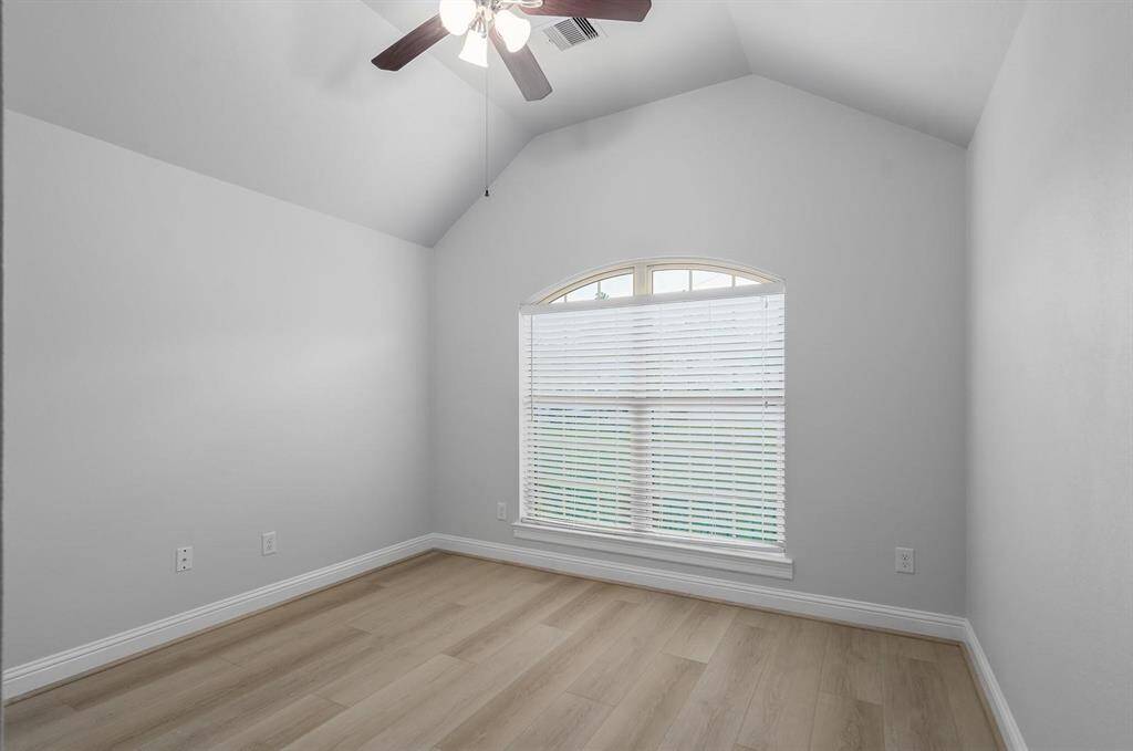 This room features a vaulted ceiling with a ceiling fan, a large arched window with blinds allowing natural light, and light-colored hardwood floors. Ideal for a cozy bedroom or home office.