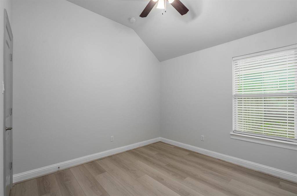 This room features a vaulted ceiling with a modern ceiling fan, light gray walls, and a window with blinds that let in natural light. It has light wood flooring, creating a clean and spacious feel.