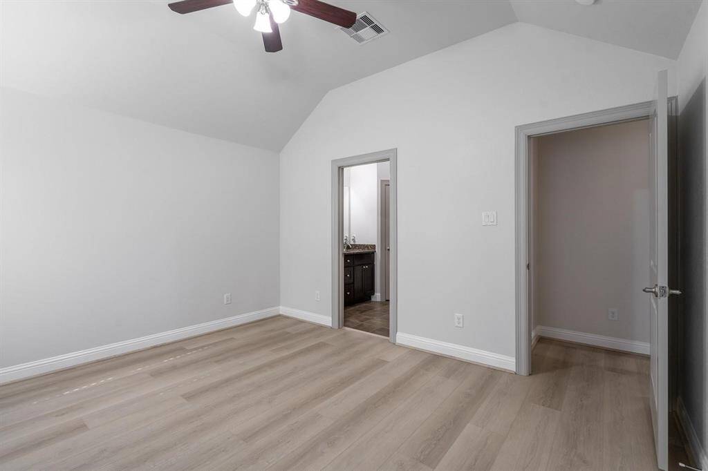 This primary bedroom features a high ceiling with a ceiling fan, light wood flooring, and neutral walls. It includes a doorway leading to a bathroom with a visible vanity and another open door leading to a separate space.