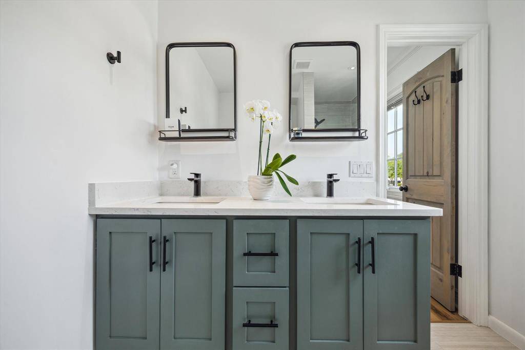 The primary bathroom features a modern double vanity with sleek black fixtures and green cabinetry. It includes two mirrors with shelves, a white countertop, and large shower. The room is bright and has a wooden door with hooks, offering a stylish and functional space