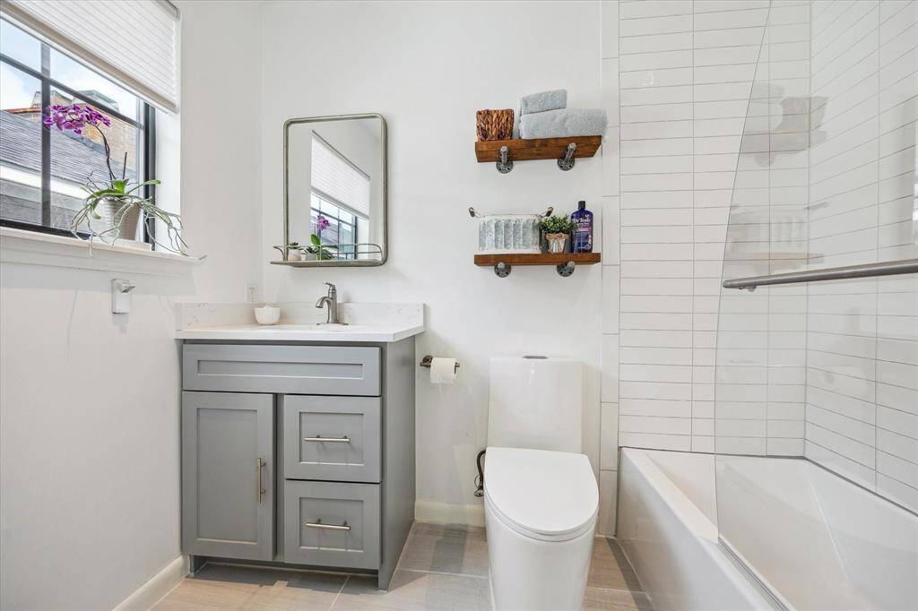 This modern bathroom features a sleek gray vanity with a white countertop, a large mirror, and open shelving for storage. The space is bright and airy, with a window providing natural light. A white tiled shower and tub with a glass panel completes the clean, contemporary look