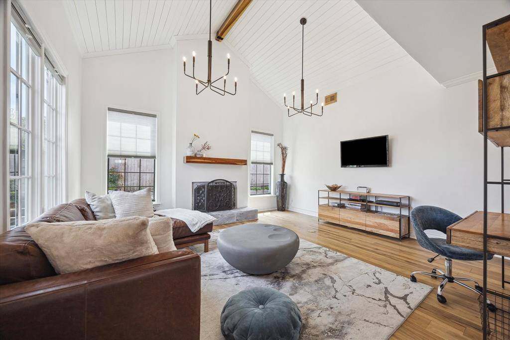 This inviting living room features high vaulted ceilings, large windows for natural light, and modern chandeliers. It includes a cozy fireplace, a sleek TV setup, and a mix of comfortable and stylish finishes, creating a perfect blend of warmth and contemporary design.