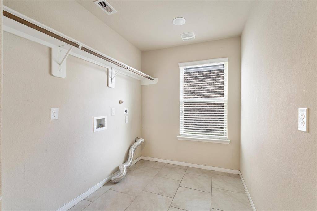 Laundry room with built-in hanging rod, utility hookups, and natural light from a large window. The space features durable tile flooring and ample room for full-size washer and dryer units.