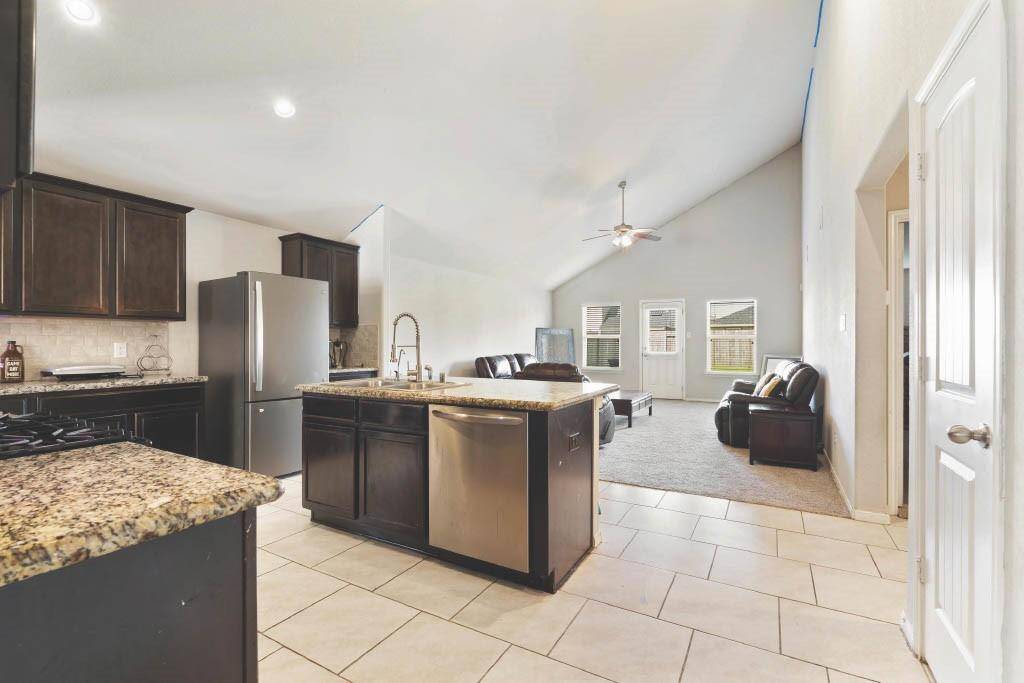 Spacious open-concept kitchen and living room featuring granite countertops, stainless steel appliances, and rich espresso cabinetry. The large center island with a deep sink and bar seating overlooks the inviting living area, highlighted by vaulted ceilings, abundant natural light, and backyard access. Perfect for everyday living and entertaining.