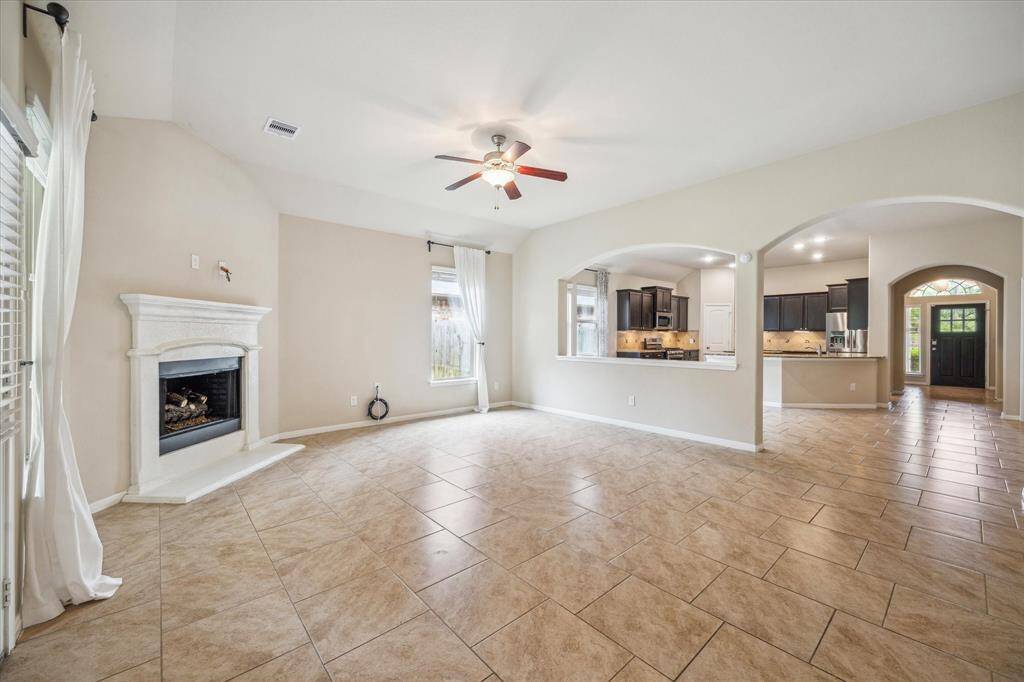 This spacious living area features a tiled floor, a cozy fireplace, and a ceiling fan. It seamlessly connects to an open kitchen with dark cabinets and stainless steel appliances, creating an inviting and functional space for family gatherings. Large windows and neutral tones enhance the bright, airy feel.