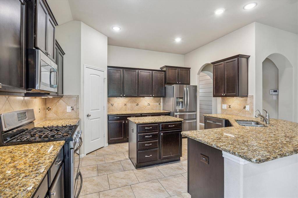 This kitchen features dark wood cabinetry, granite countertops, a central island, and stainless steel appliances. It offers a spacious layout with tiled flooring and recessed lighting, perfect for modern living.