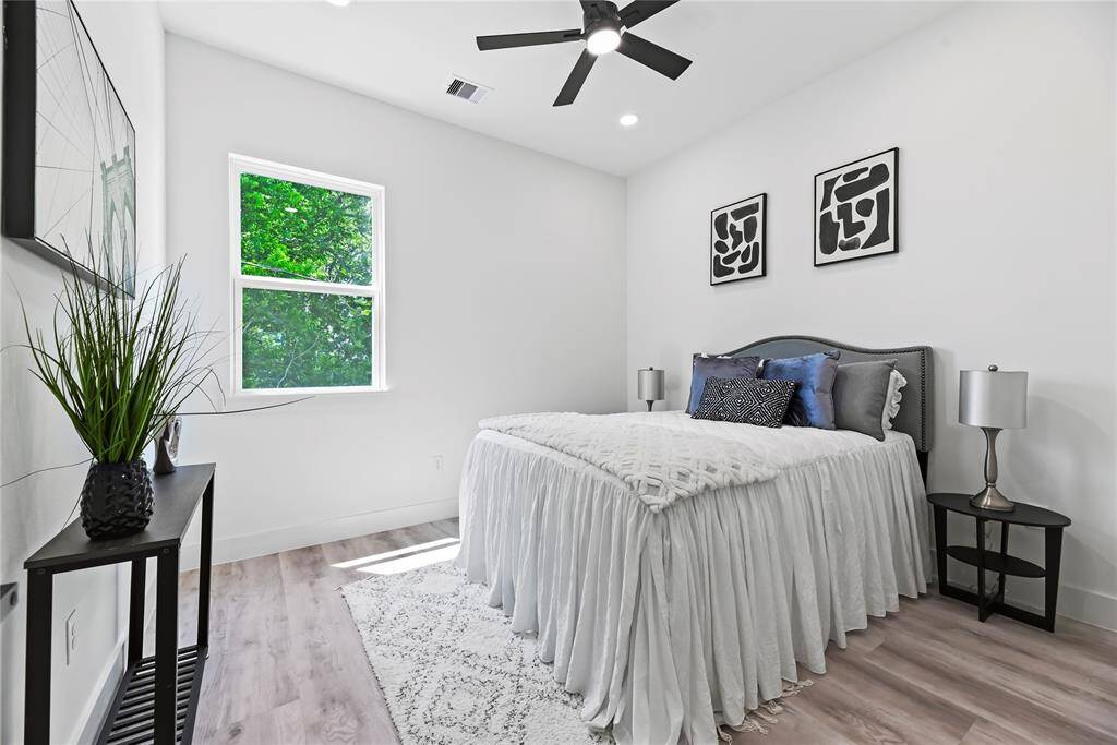 This bright bedroom features a modern design with white walls. It includes a ceiling fan and natural light from a large window, creating a calming and inviting atmosphere.