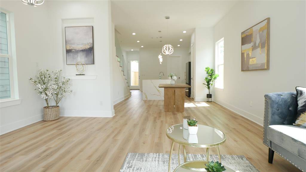 This photo showcases a bright, open-concept living space with modern decor. It features light wood flooring, a cozy seating area, and a view into the kitchen with stylish pendant lighting. Large windows allow natural light to illuminate the space, creating an inviting atmosphere.