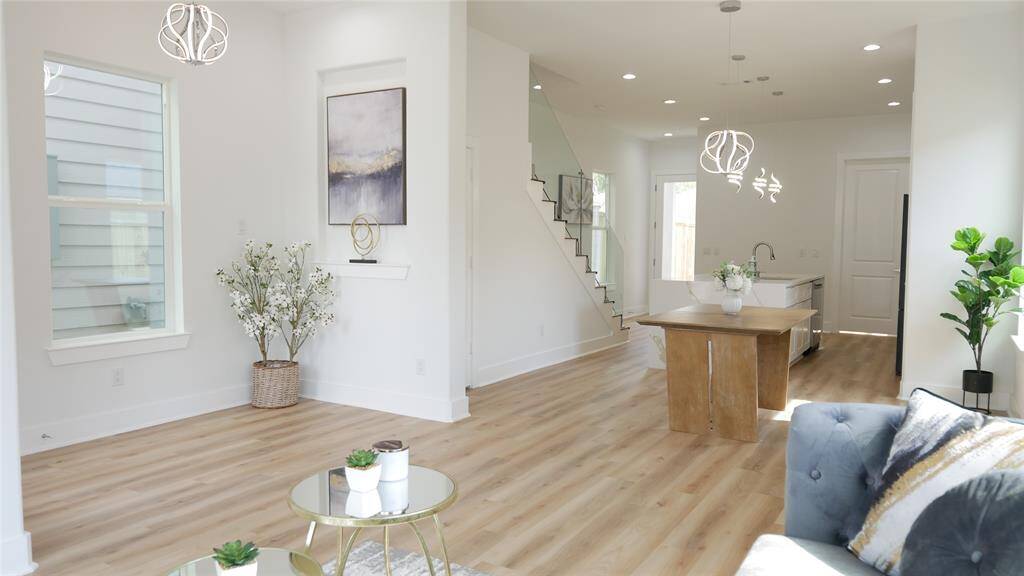 Bright and modern open-concept living area with light wood flooring and elegant lighting fixtures. Features a stylish kitchen island, contemporary staircase, and large windows for natural light. Ideal for comfortable living and entertaining.