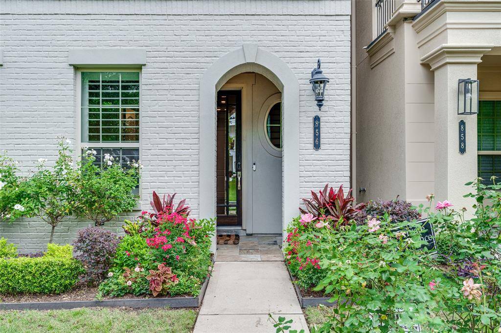 Elegant entryway with a light brick exterior and arched doorway, surrounded by lush landscaping and vibrant flowers.