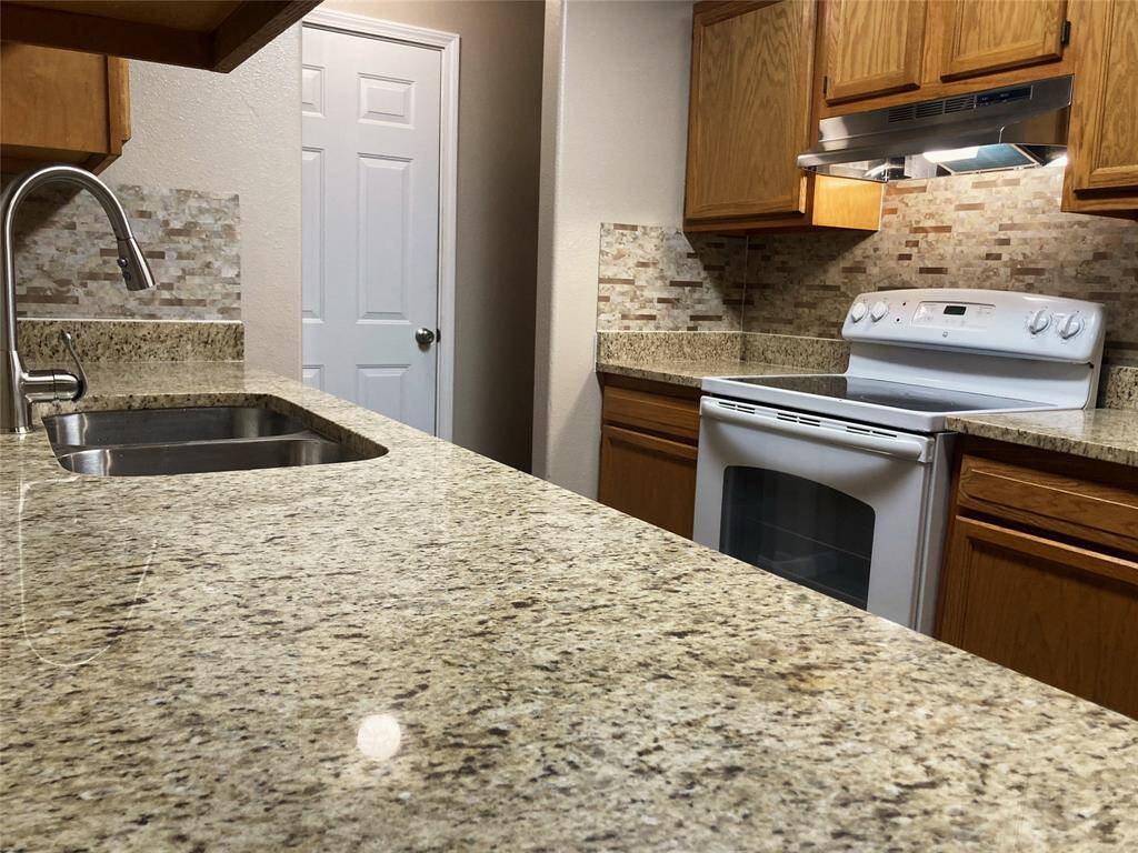 This kitchen features granite countertops, a double sink, and a modern electric stove. It has wooden cabinets and a neutral backsplash for a warm, classic look.