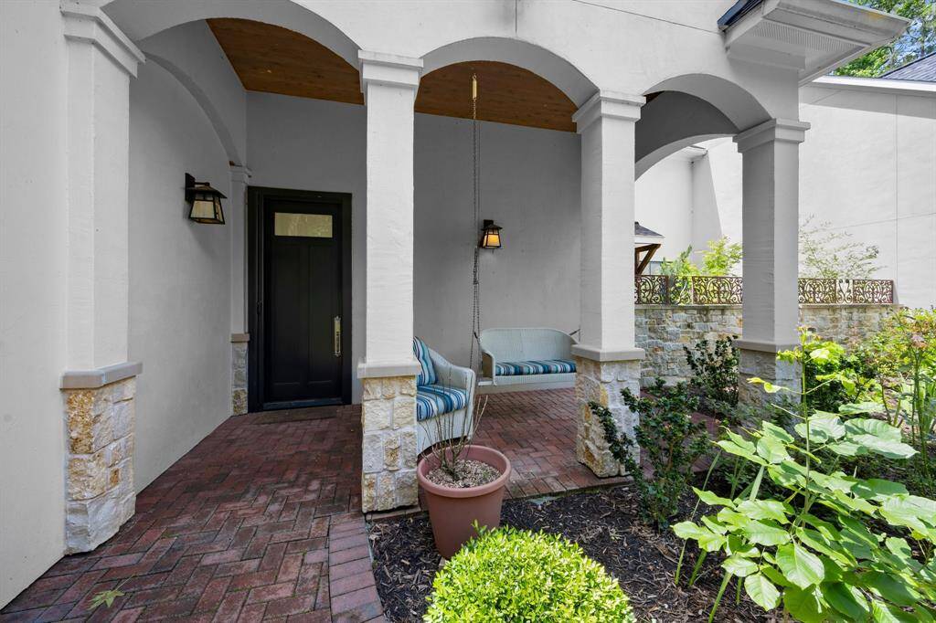 This inviting home entrance features a charming covered porch with archways, brick flooring, and cozy seating. Surrounded by lush greenery, it offers a warm and welcoming atmosphere.