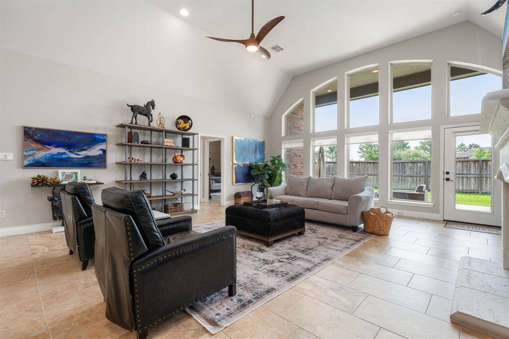 This stunning living area showcases soaring vaulted ceilings, a wall of expansive windows that flood the space with natural light and elegant tile flooring throughout. The open and airy layout is perfect for both relaxing and entertaining with stylish finishes and a seamless indoor-outdoor connection through the glass door leading to the backyard.