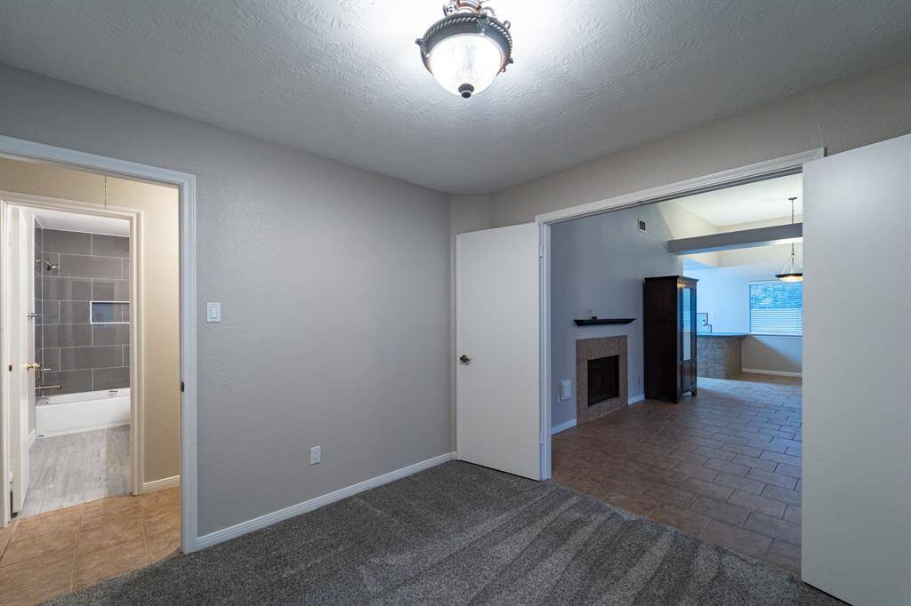 This photo features a cozy room with carpet flooring leading into a tiled living area. There's a fireplace and ample natural light from a large window. An en suite bathroom, with modern tile work, is visible in the background.