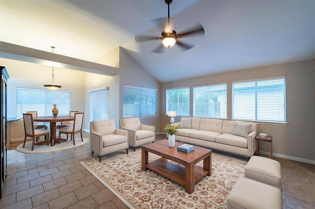 Bright and spacious living area featuring a vaulted ceiling, tile flooring, and ample natural light from large windows. The room includes a comfortable seating arrangement with a neutral palette, a ceiling fan, and a cozy dining nook with a round table.
