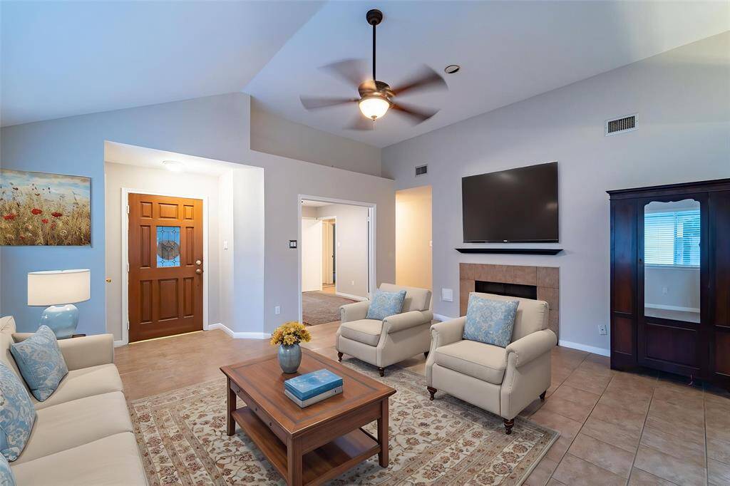 This inviting living room features a high ceiling with a ceiling fan, a cozy fireplace, and a mounted TV. It has tile flooring, a neutral color scheme, and comfortable seating, creating a warm and welcoming atmosphere. A wooden door with decorative glass adds charm.