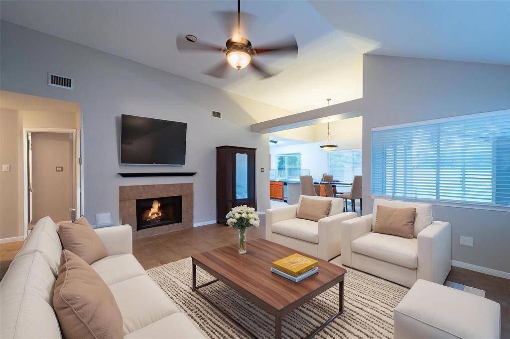 This inviting living room features a cozy fireplace, modern ceiling fan, and ample natural light. It opens to a dining area, perfect for entertaining. Neutral tones and comfortable seating create a warm atmosphere.