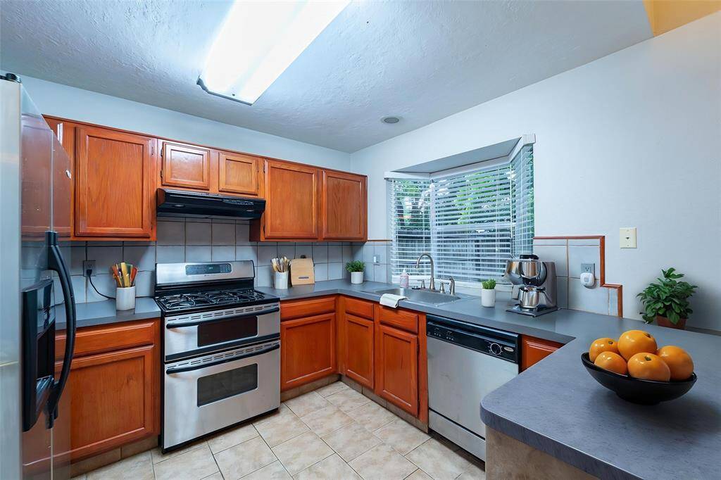 This kitchen features warm wooden cabinetry, stainless steel appliances, and a spacious countertop. A large window provides natural light, and the tile flooring adds a practical touch. Perfect for cooking and entertaining.