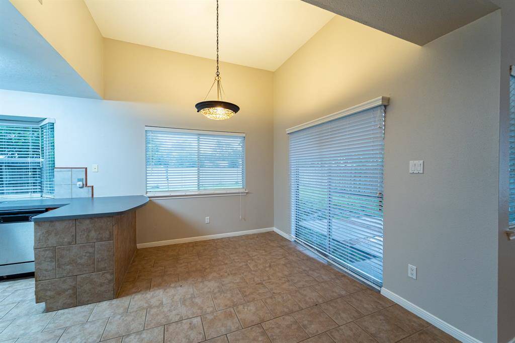 This room features a high ceiling with a modern light fixture, large windows with blinds, and a sliding glass door leading to a patio. The space connects to a kitchen area with a tiled bar counter, offering an open and bright layout.