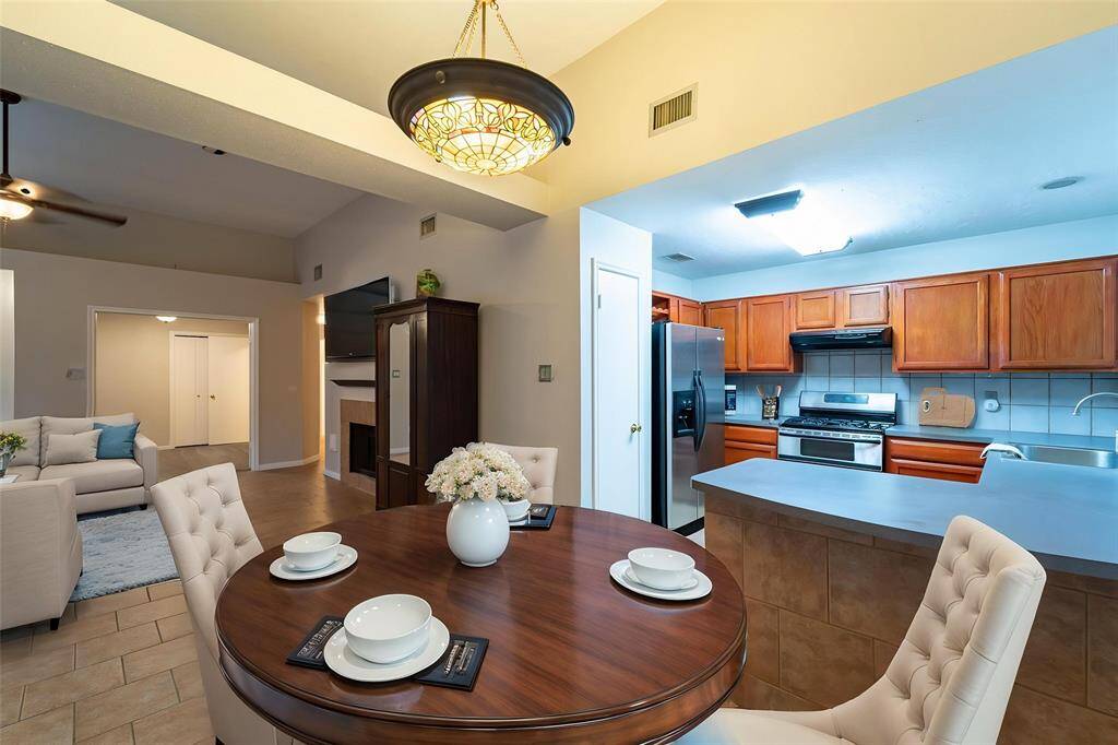 This photo showcases a cozy, open-concept living area with a modern kitchen featuring wooden cabinets and stainless steel appliances. There's a dining area with elegant seating and a pendant light, while the adjacent living room offers a comfortable space with neutral decor.