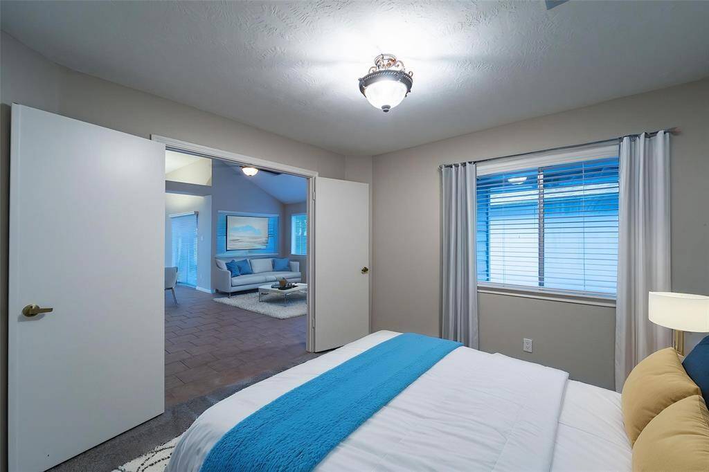 This photo shows a cozy bedroom with neutral walls and a ceiling light fixture. It features a window with blinds and curtains, and an open door leading to a spacious living area with natural light and modern furnishings.