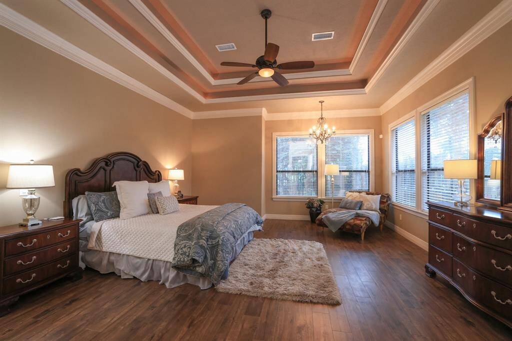 This spacious bedroom features a luxurious tray ceiling with crown molding, rich luxury vinyl plank flooring, and ample natural light from large windows.
