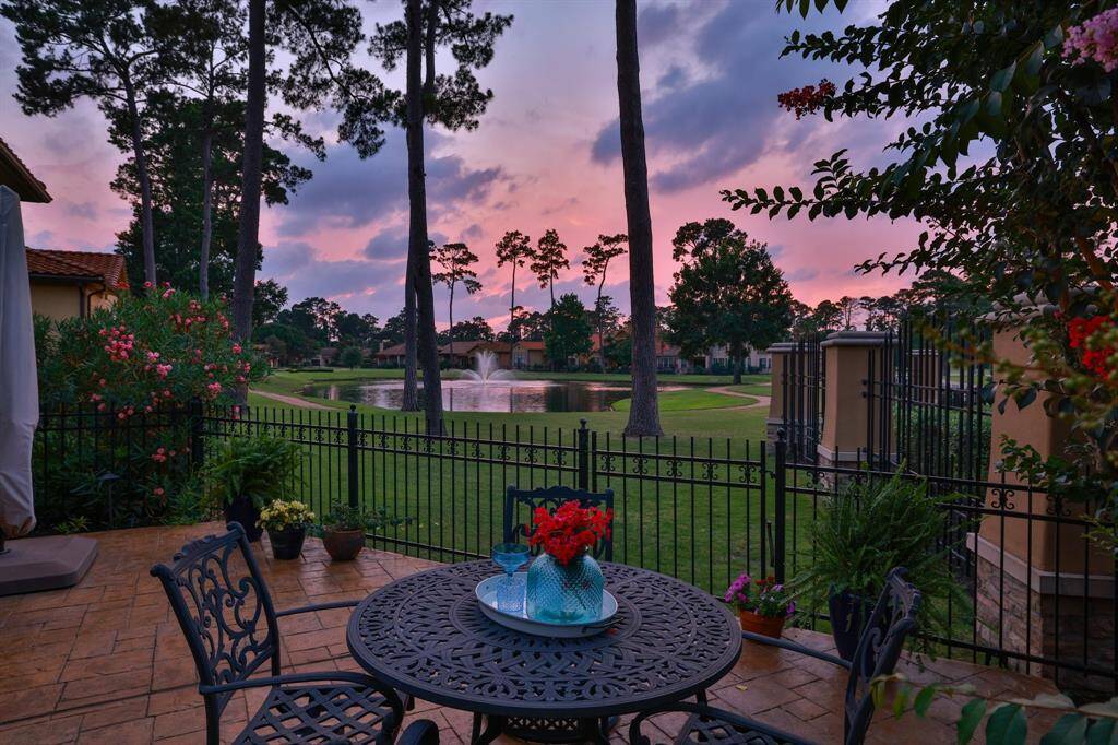 This inviting patio offers a serene view of a neighborhood  pond with a fountain, surrounded by tall trees. The space features comfortable seating and a dining area, perfect for relaxing or entertaining while enjoying picturesque sunsets.
