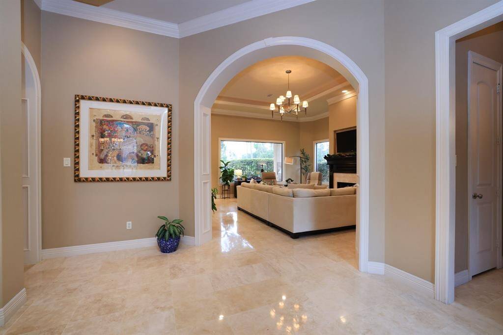 This photo showcases a spacious, elegant living area with a warm, neutral color palette. The room features polished Travertine tile flooring, a large archway leading to a cozy sitting area, and a stylish chandelier.