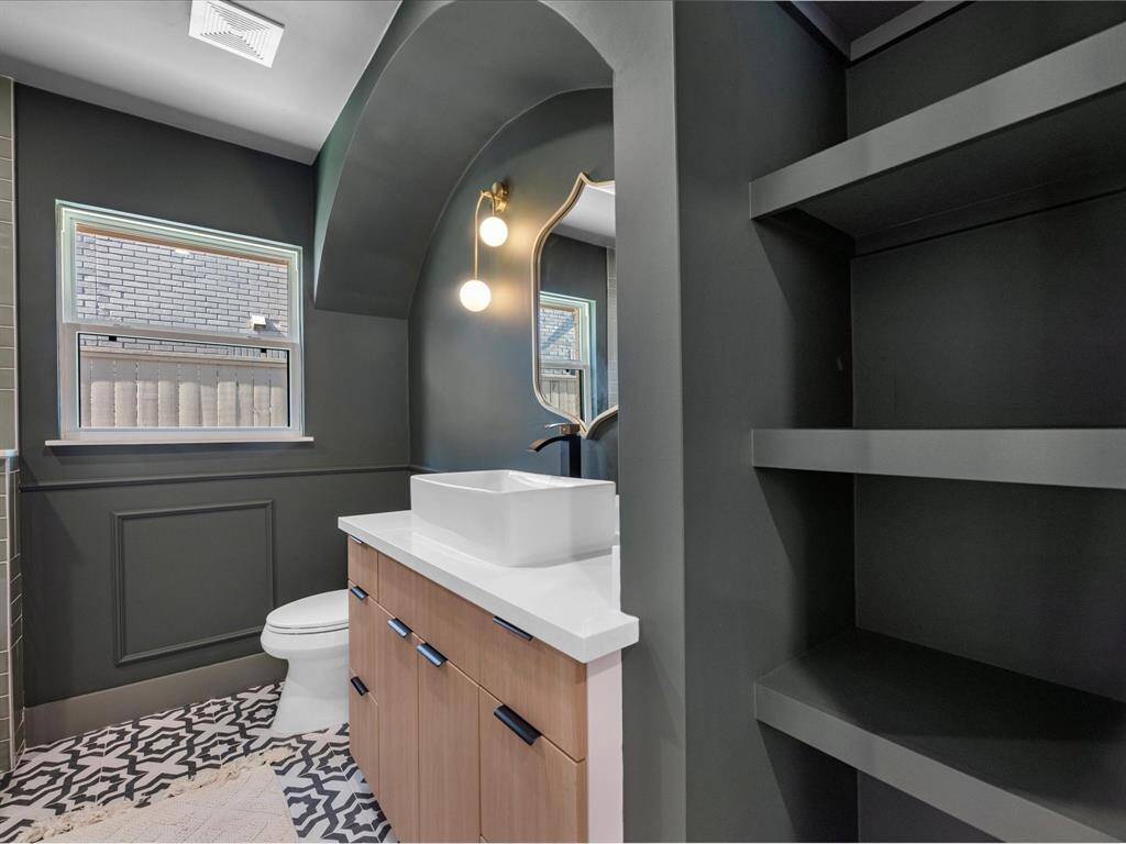 The first-floor hallway bathroom features bold, modern design with arched accents, custom shelving, a vessel sink with quartz countertop, designer lighting, and patterned floor tiles for a striking yet functional space.