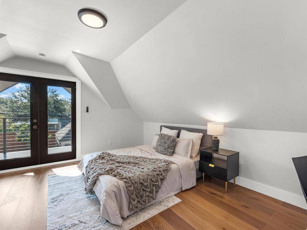 The upstairs third bedroom features a cozy design with sloped ceilings, rich wood flooring, and private balcony access through elegant French doors, offering serene views and natural light.