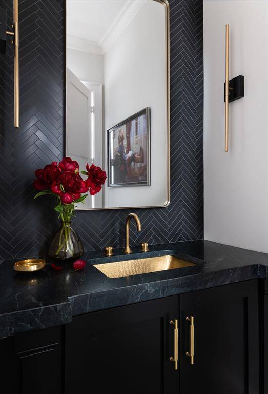 A designer powder room exudes elegance with its stunning color palette, complemented by a brass sink and fixtures. The luxurious stone countertop and herringbone tile that stretches to the ceiling create a striking focal point.