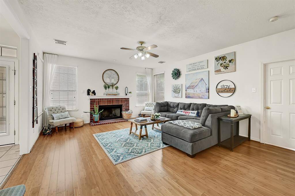 Inviting living room with bamboo flooring and a cozy brick fireplace, perfect for relaxing or entertaining. Large windows bring in natural light, and the open layout flows effortlessly into the kitchen and dining area.