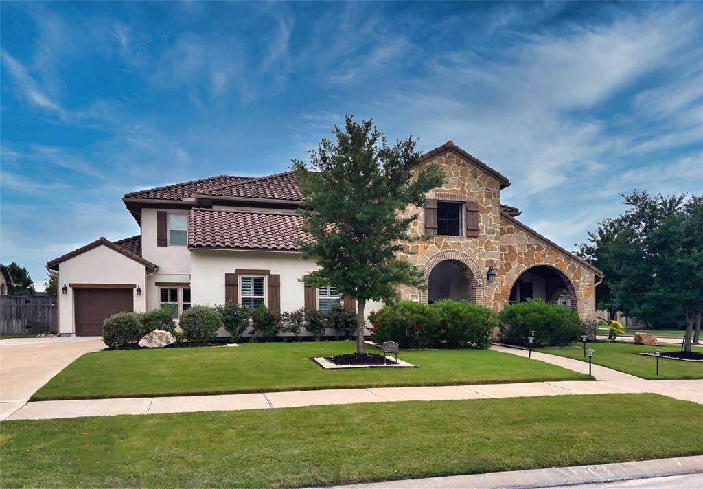 This is a beautiful two-story home with a mix of stone and stucco exterior, featuring a tiled roof and spacious arched entryway. It has a well-maintained front yard with lush greenery and a spacious driveway leading to the garages.