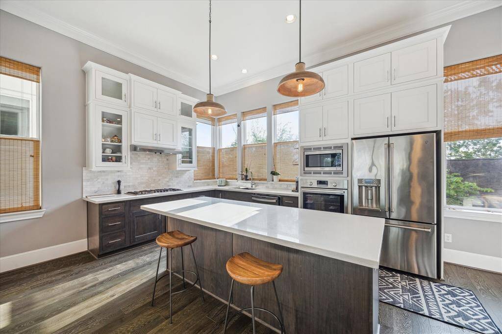 This modern kitchen features high ceilings, a large island with seating, stainless steel appliances, and ample natural light from multiple windows. The space combines sleek white cabinetry with dark wood accents and stylish pendant lighting.