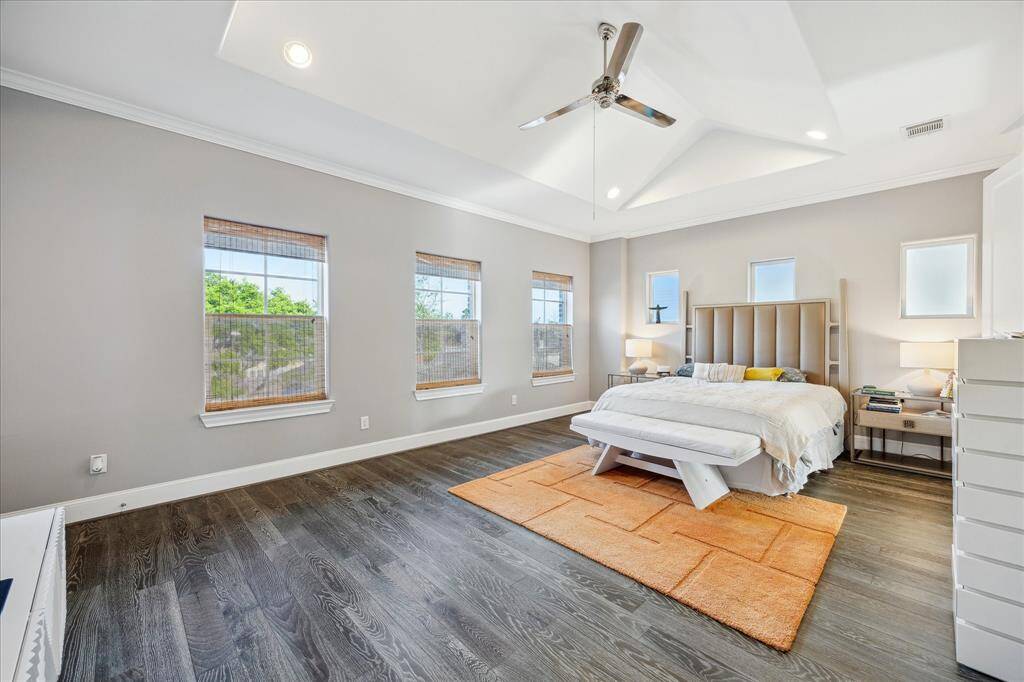 Primary bedroom with high ceilings, featuring large windows for natural light, sleek dark wood flooring, and a stylish ceiling fan. The neutral color palette and elegant furnishings create a calming atmosphere.