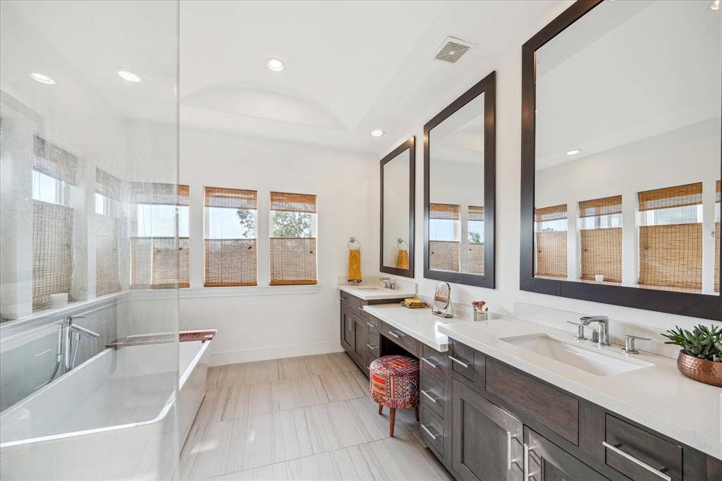 Primary bathroom features a modern design with dual sinks, a spacious vanity, large mirrors, and a soaking tub. Natural light streams through multiple windows with stylish bamboo shades, creating a bright and inviting space.