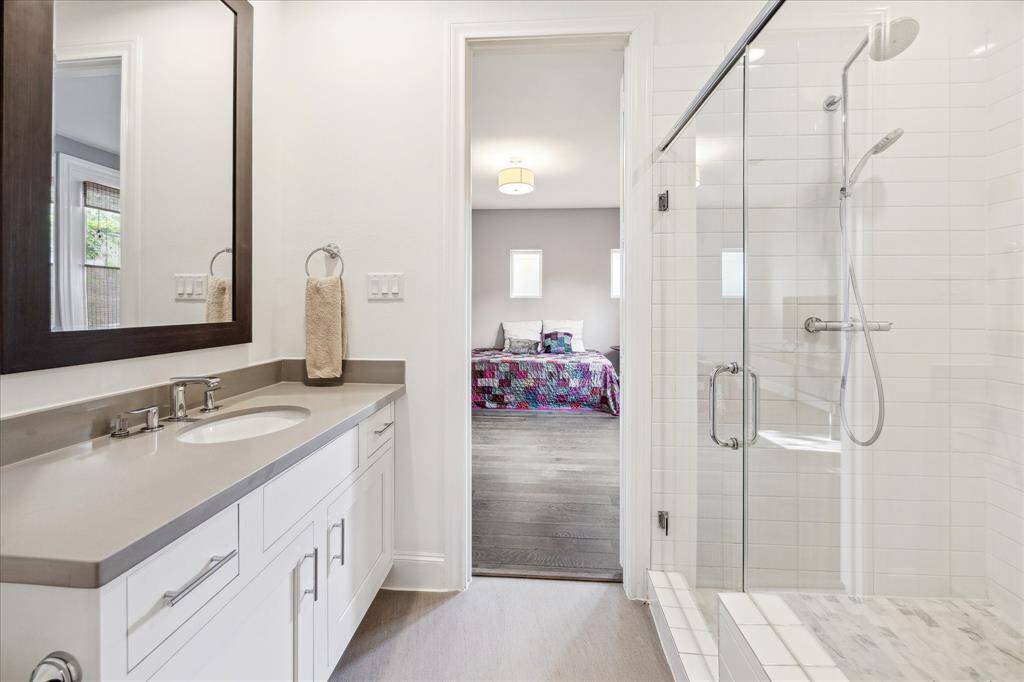 Secondary bathroom with a sleek vanity and large mirror on the left, a glass-enclosed shower on the right, and an adjoining view into a bedroom featuring a bed with colorful bedding and natural light.