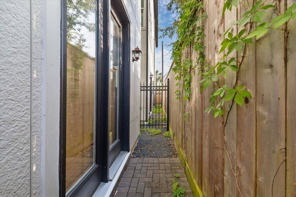 Side pathway with a wooden fence on one side and a house wall with large windows on the other. It features a brick walkway, some greenery, and a small gate at the end.