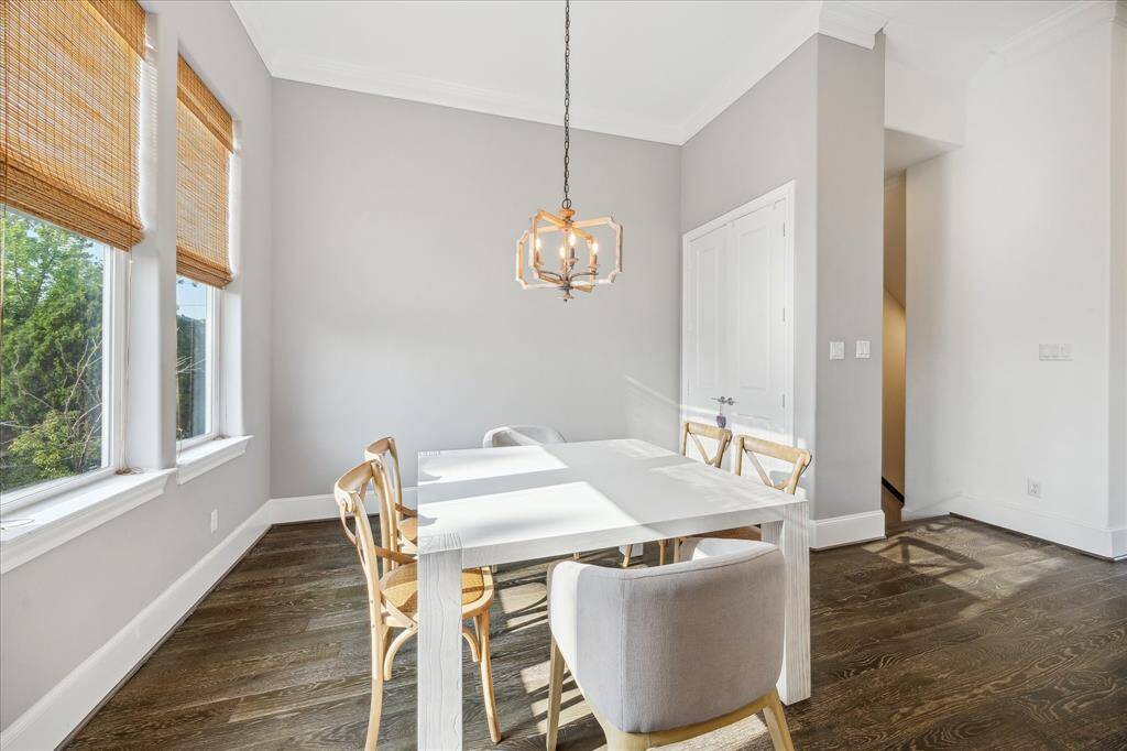 Bright dining area with large windows, attractive chandelier, and neutral tones. Features a sleek white table and wooden chairs on dark hardwood floors, creating a cozy yet elegant space.
