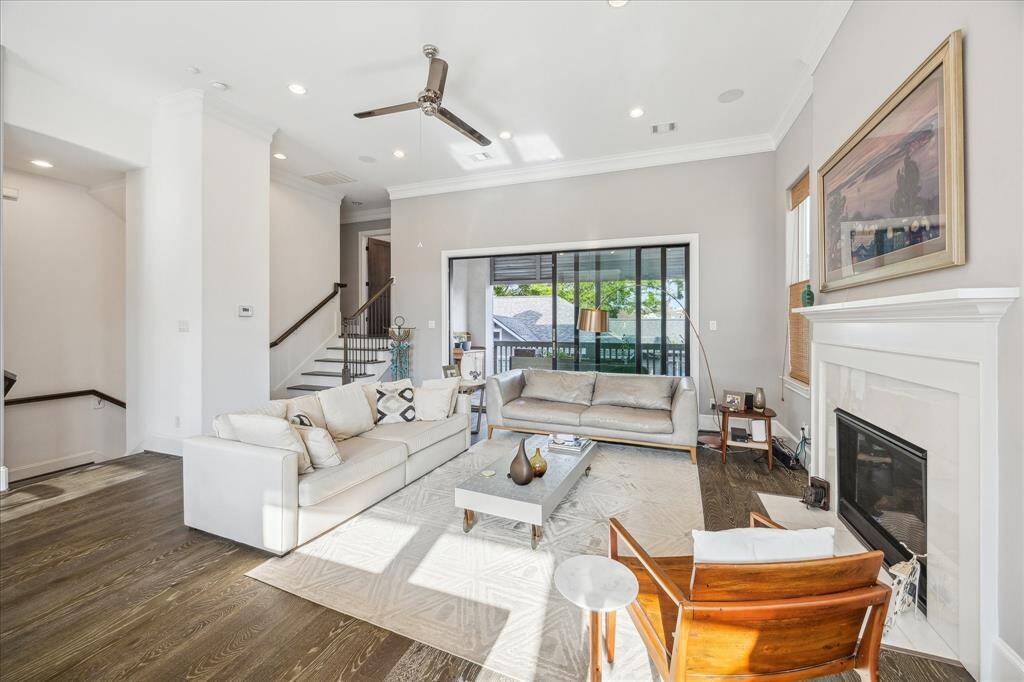 This spacious living room features high ceilings, a modern ceiling fan, and abundant natural light. It includes a cozy fireplace, sleek hardwood floors, and a blend of contemporary and classic furnishings. Large windows and a sliding glass door provide access to a balcony, enhancing the open and airy feel.