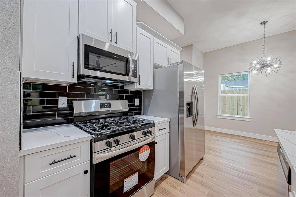 This photo showcases a modern kitchen with stainless steel appliances, including a gas range and refrigerator. It features white cabinetry, a sleek black tile backsplash, and light wood flooring. A stylish pendant light and a window with a view complete the space.