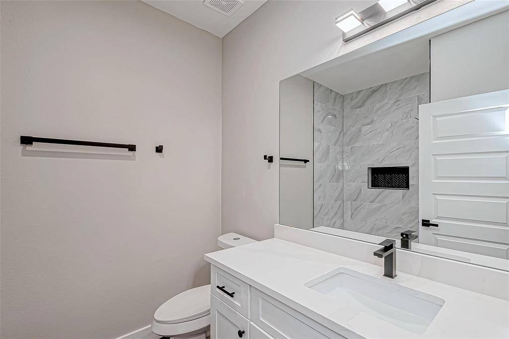 This modern bathroom features sleek white cabinetry, a large mirror, and a stylish black faucet. The spacious shower has elegant tile work with a built-in niche for storage. The minimalist design provides a clean and contemporary feel.