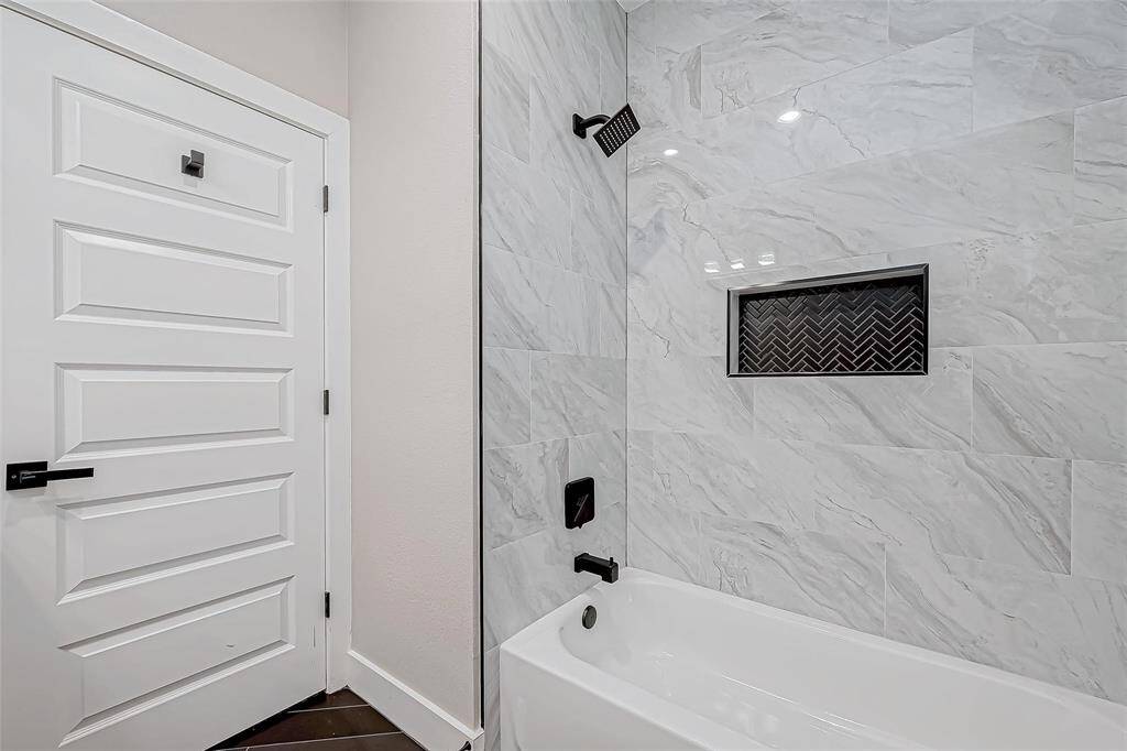 This bathroom features a sleek tub with modern black fixtures, a white marble tile surround, and a stylish inset shelf with a herringbone pattern. The bright, clean design is complemented by a paneled white door.