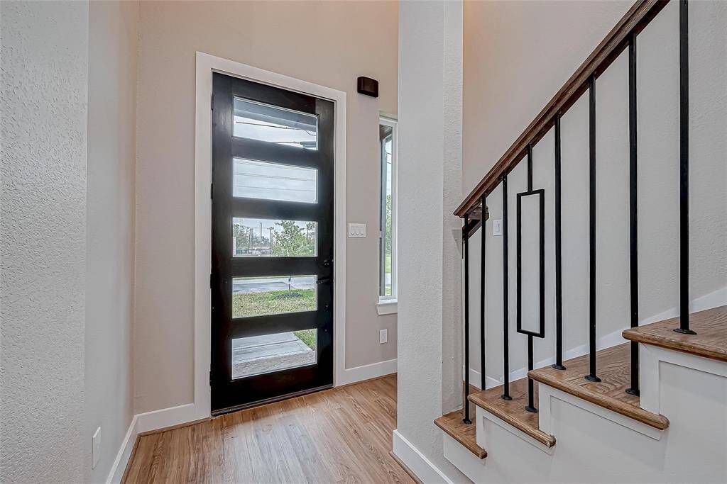 This entryway features a modern black front door with frosted glass panels, wood flooring, and a staircase with sleek black metal railings. There’s a side window for natural light, creating an inviting atmosphere.