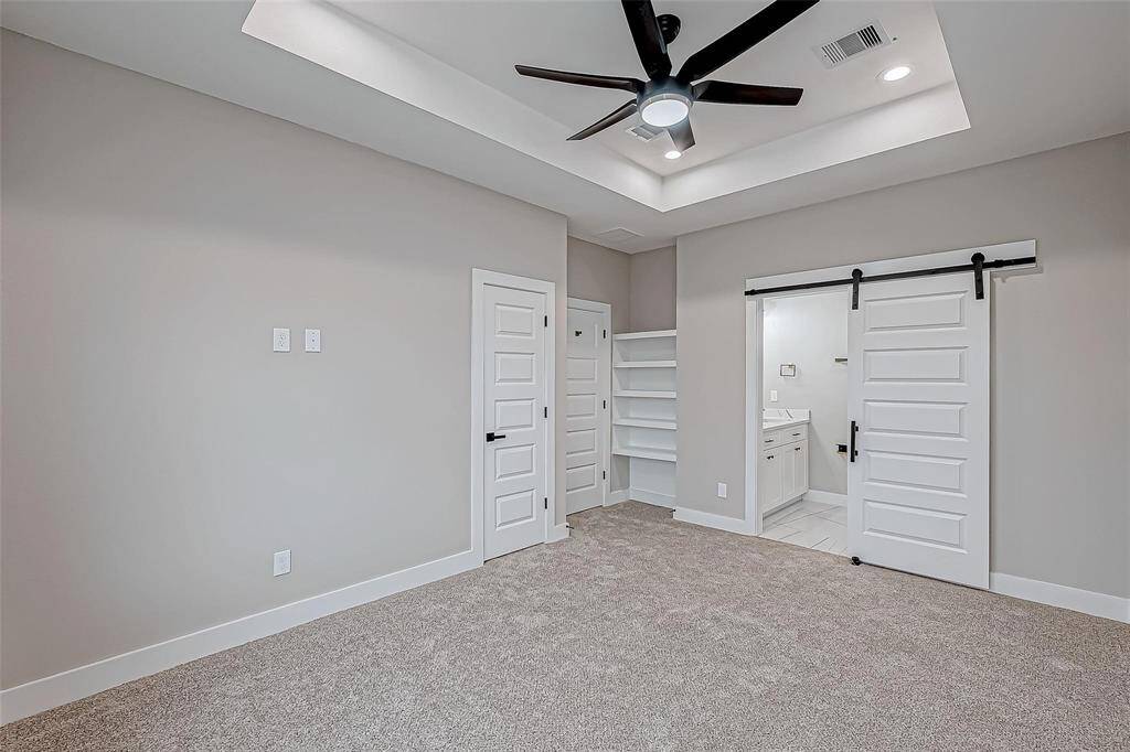 This photo features a modern master-bedroom with soft gray walls and plush carpet. It includes a ceiling fan, recessed lighting, and a tray ceiling for added elegance. There's a sliding barn door leading to an adjoining bathroom, and built-in shelving offers extra storage space.