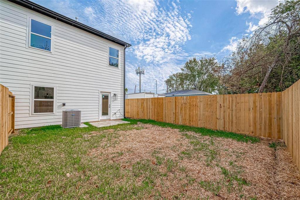 This photo shows a fenced backyard with a grassy area and a two-story house exterior. There’s an air conditioning unit next to the back door, and the space is framed by wooden fencing, providing privacy.