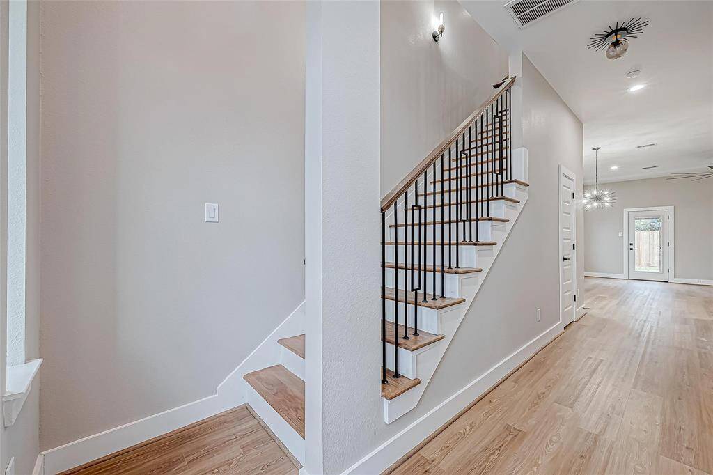 This photo showcases a modern, open-concept entryway with light wood flooring. A stylish staircase with wooden steps and black metal railing leads upstairs. Adjacent is a bright hallway leading to a room with pendant lighting and a door to the backyard.